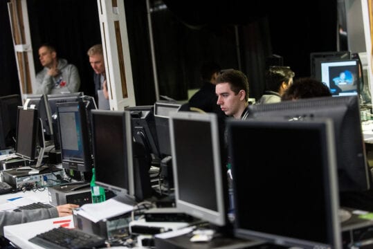 Computers lined up on a long office desk and some IT operators.